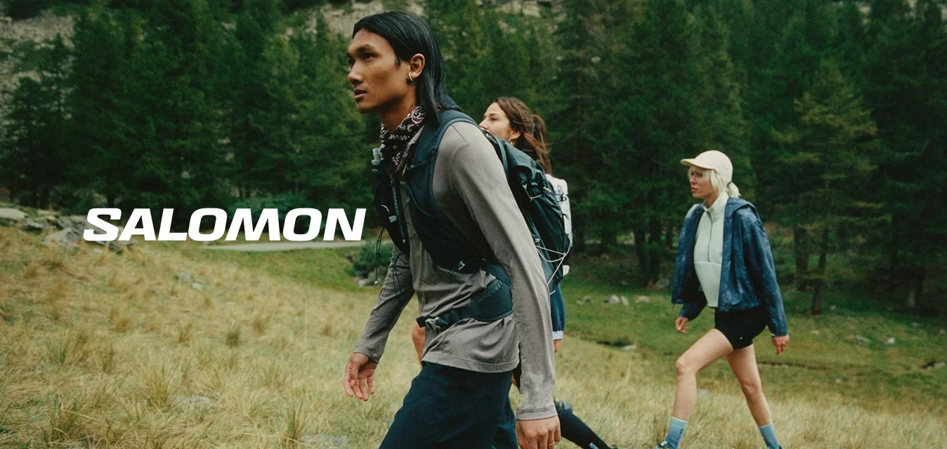 Tre individui camminano su un terreno erboso. Indossano abbigliamento outdoor: giacche leggere, pantaloni corti e zaini. Il contesto presenta alberi e rocce.