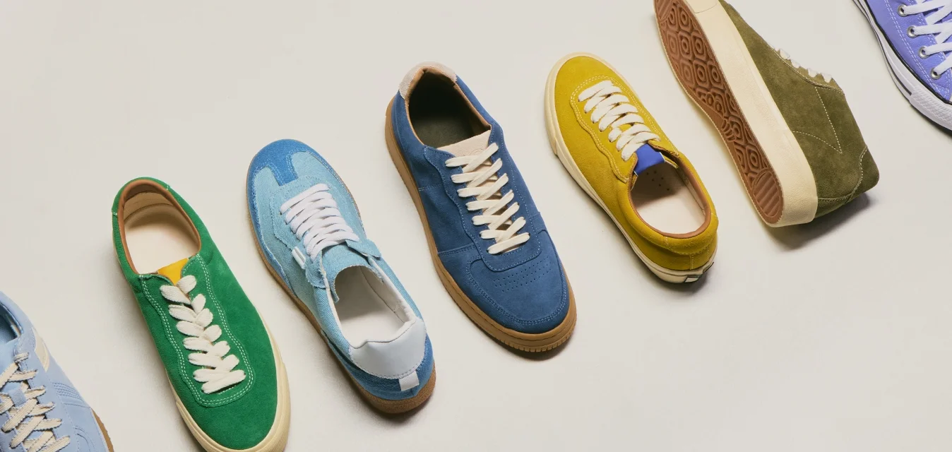 Sette sneaker casual in suede di vari colori, tra cui blu, verde, giallo e viola, disposte in fila su uno sfondo chiaro.