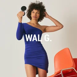 Donna con un vestito blu monospalla tiene uno specchio compatto rotondo, in piedi accanto a una pila di quattro sedie arancioni su uno sfondo semplice.