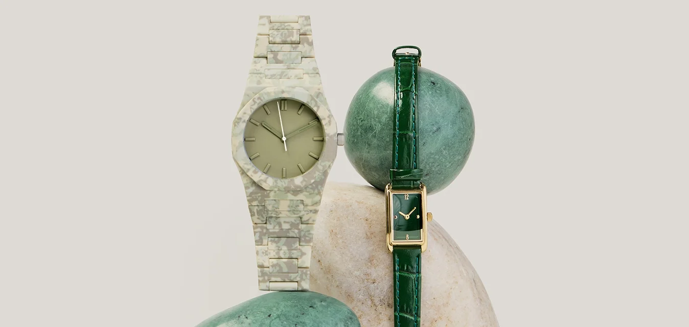 Due orologi da polso, uno con cinturino in pelle verde e quadrante rettangolare, l'altro con cinturino mimetico e quadrante rotondo, esposti su delle pietre.