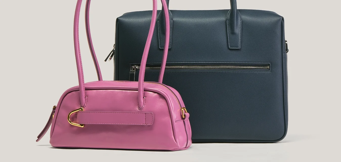 Piccola borsa rosa con manici lunghi accanto a una borsa più grande stile ventiquattrore blu scuro con tasca frontale con zip su sfondo semplice.