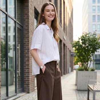 Smiling young woman standing on a city pavement wearing a white shirt and brown trousers, casual urban fashion