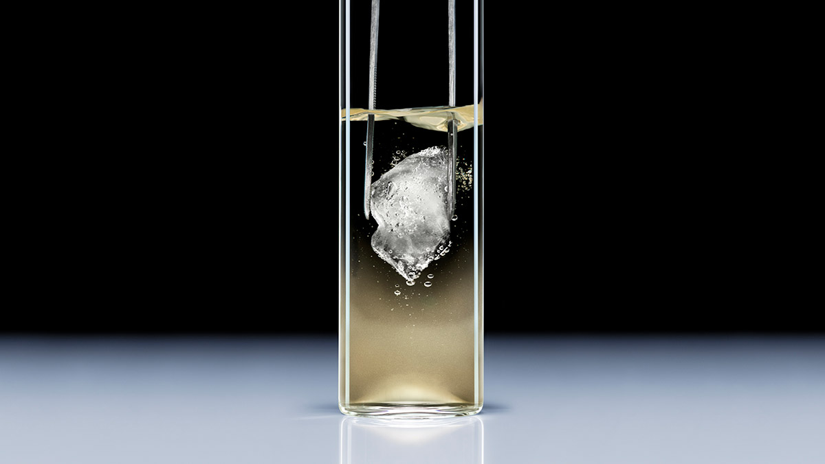 Un cubetto di ghiaccio trattenuto da una pinza e immerso in un elisir dorato, che illustra l'effetto criogenico rinfrescante della formula Replasty. 