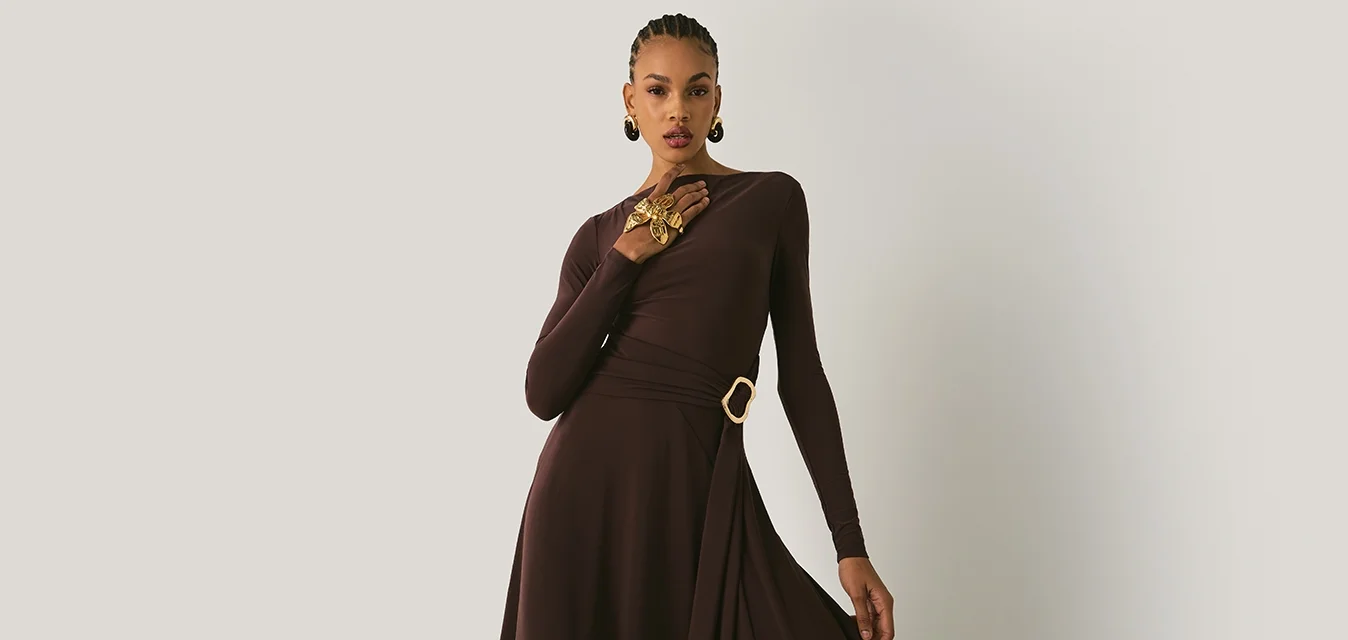 Abito lungo marrone con maniche a lunghezza completa e design avvolgente, caratterizzato da una fascia laterale e un accento a forma di anello dorato in vita. Tessuto morbido con una silhouette fluida.