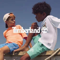 Due ragazzi sono seduti su un molo di legno sopra l'acqua, uno tiene una corda, sotto un cielo azzurro limpido con alberi lontani.