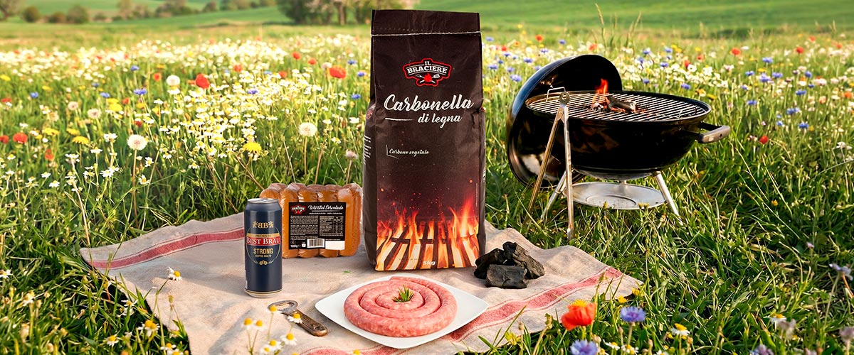 Barbecue setup in a wildflower meadow: bag of charcoal, lit kettle grill, raw coiled sausage on a plate, sausage pack and beer can on a picnic cloth