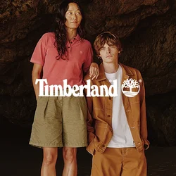 Due persone sono in piedi sulla sabbia vicino all'ingresso di una grotta rocciosa; una è sopra una roccia, indossa una camicia rosa e pantaloncini, l'altra una giacca marrone e pantaloncini.