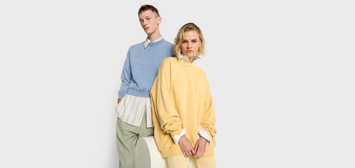 Due modelli indossano abbigliamento casual: uno in un maglione azzurro chiaro sopra una camicia bianca a fantasia, e l'altro in una felpa gialla con pantaloni beige.