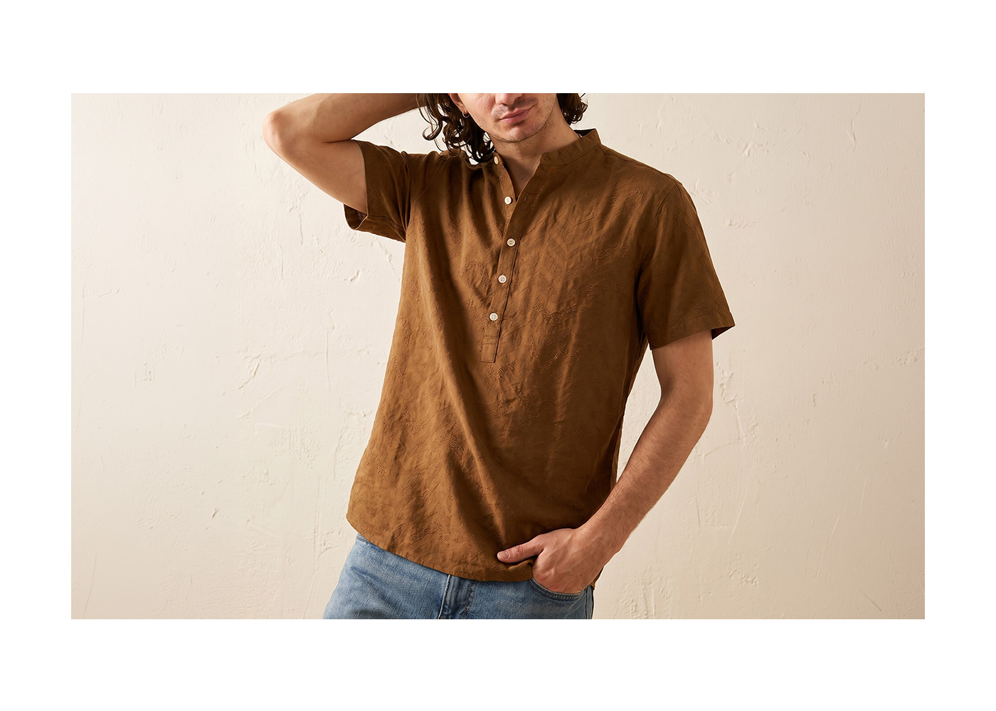 Man wearing a brown short-sleeve linen shirt with mandarin collar and button placket, styled with blue jeans against a textured neutral wall