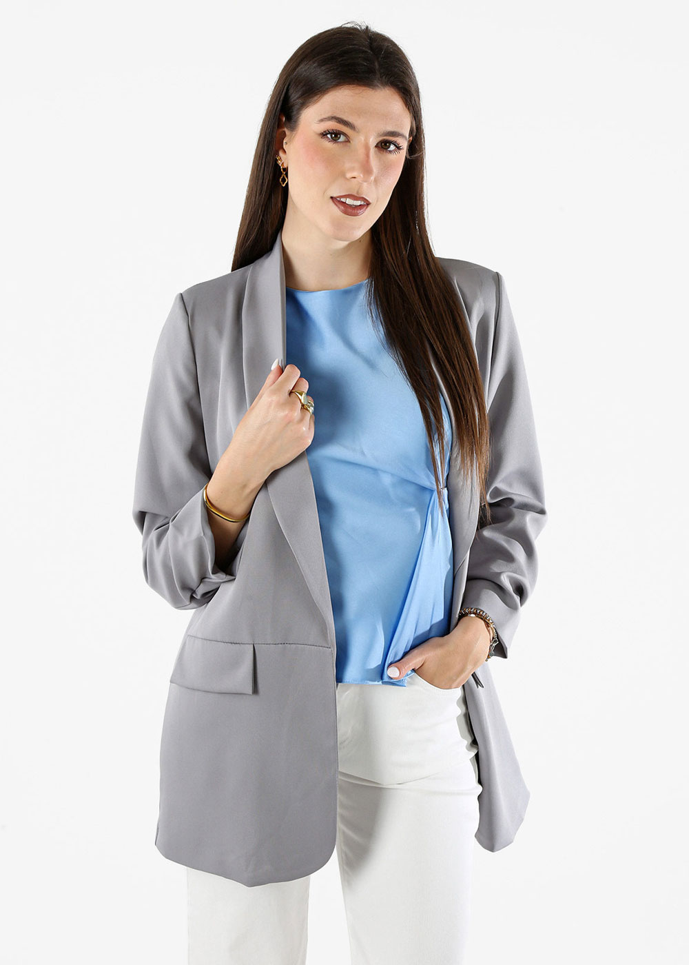 Woman wearing a grey blazer, blue blouse and white trousers, standing with hand in pocket and long brown hair against white background