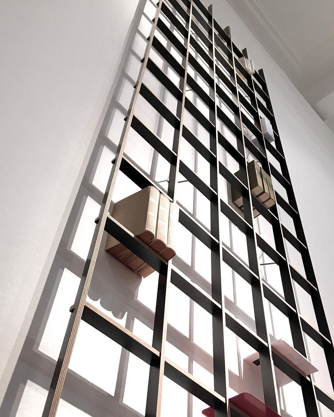 Tall wall-mounted grid bookshelf with scattered books and dramatic geometric shadows, photographed from a low upward perspective
