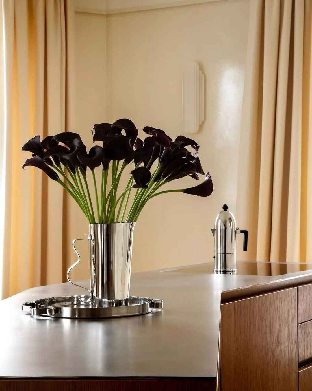 Dark purple calla lilies in a polished silver vase on a modern kitchen island beside a stainless steel coffee maker and cream curtains