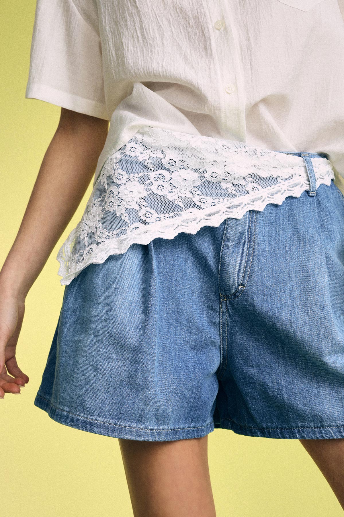 Close-up of a torso wearing a white blouse with lace trim tucked into high-waisted blue denim shorts on a yellow background