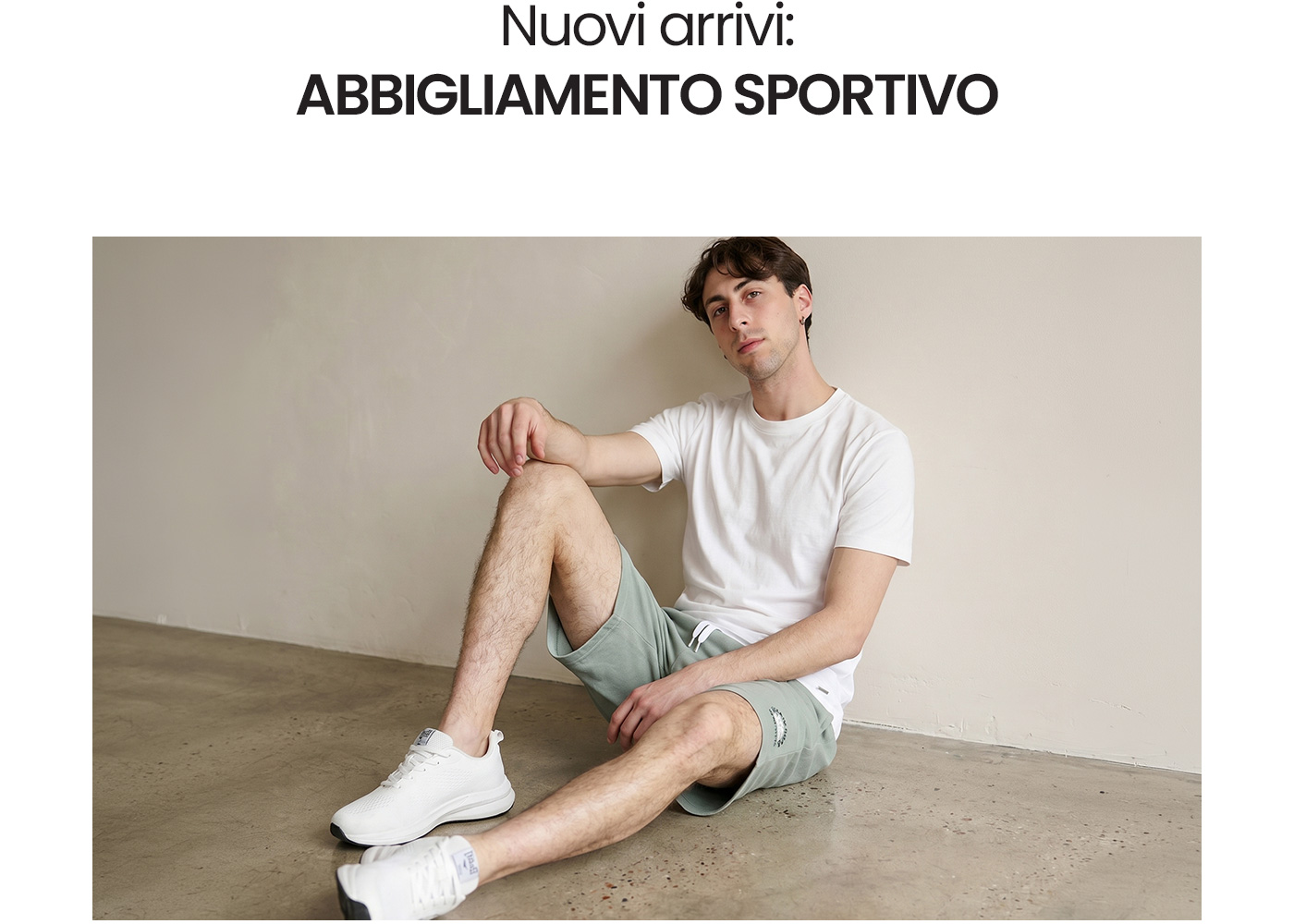Male model in white T-shirt, light green sports shorts and white trainers sitting on a concrete floor against a pale wall, showcasing sportswear