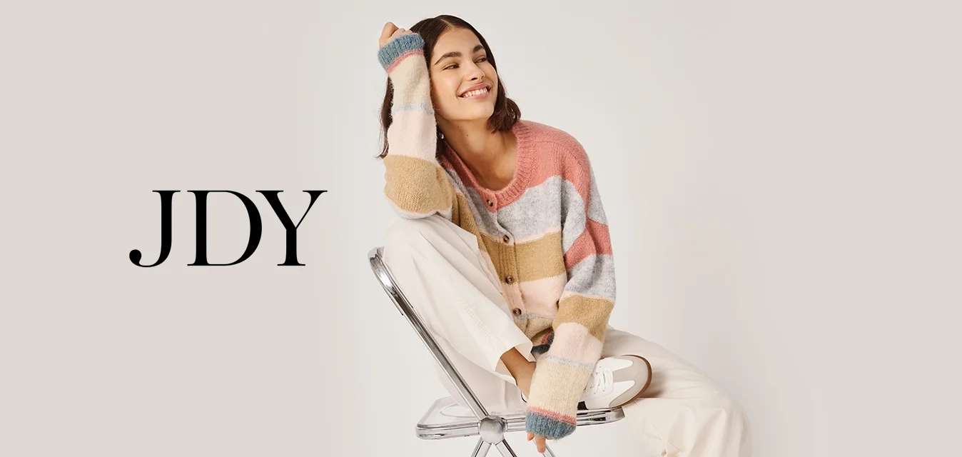 Giovane donna sorridente, seduta di lato su una sedia pieghevole in metallo, che indossa un maglione a righe multicolore, pantaloni bianchi e sneakers.