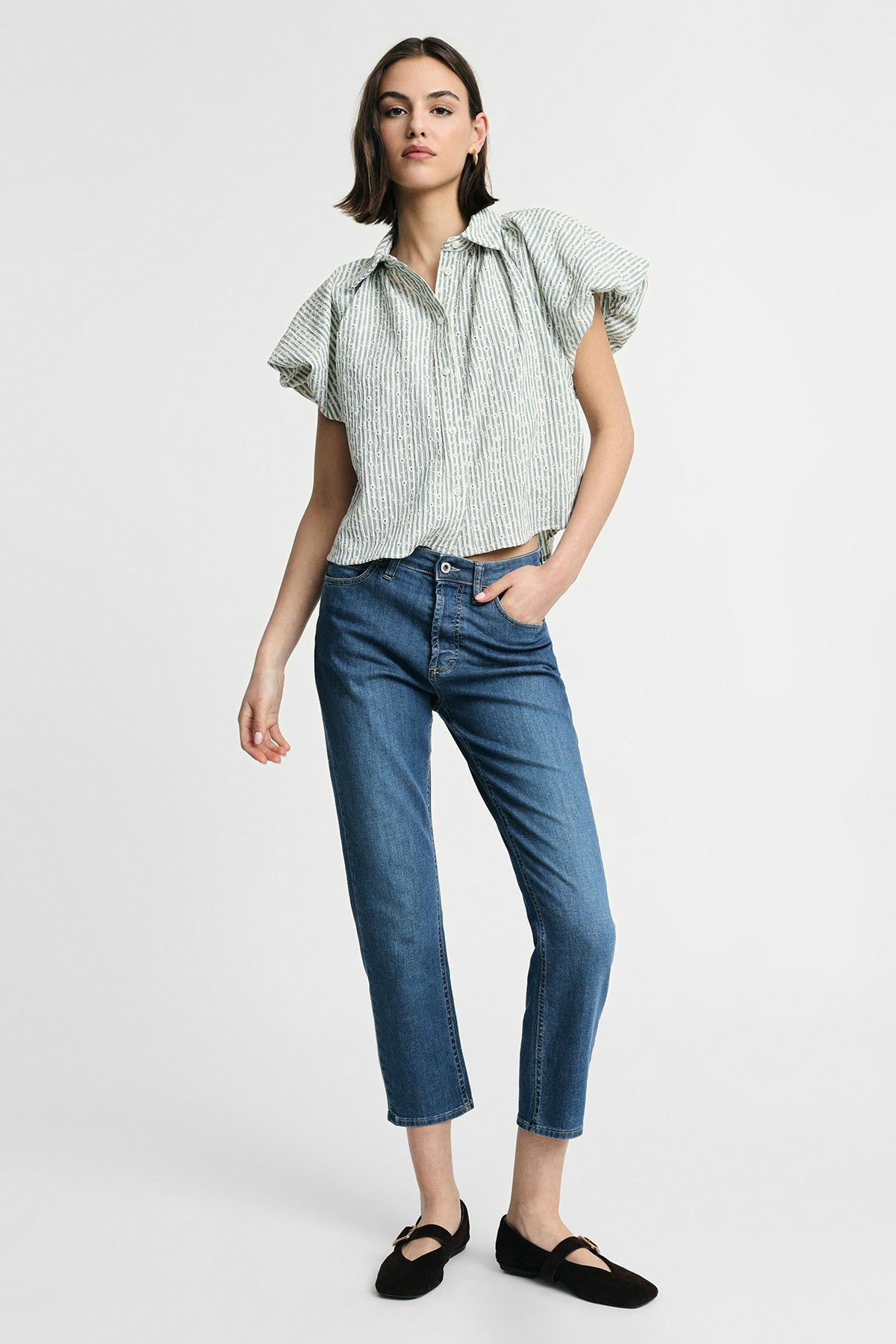 Model in green-striped short-sleeve blouse, cropped blue jeans and black ballet flats standing on a white background