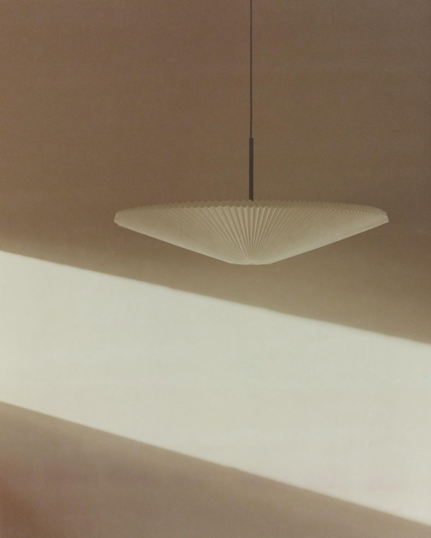 Minimalist pleated pendant lamp with conical ribbed shade suspended against a beige wall, bathed in soft diagonal sunlight and shadows