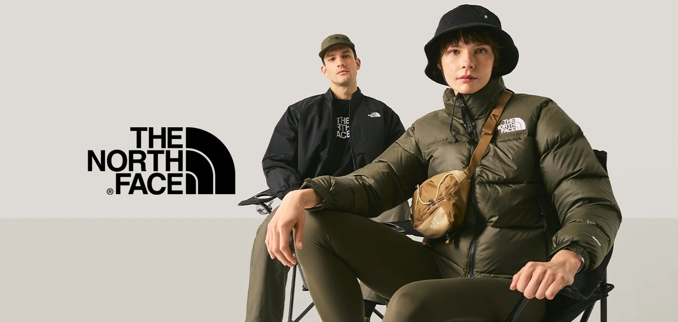 Due giovani seduti su sedie pieghevoli contro uno sfondo chiaro e semplice, uno dei quali indossa un cappello a secchiello nero e una giacca imbottita.