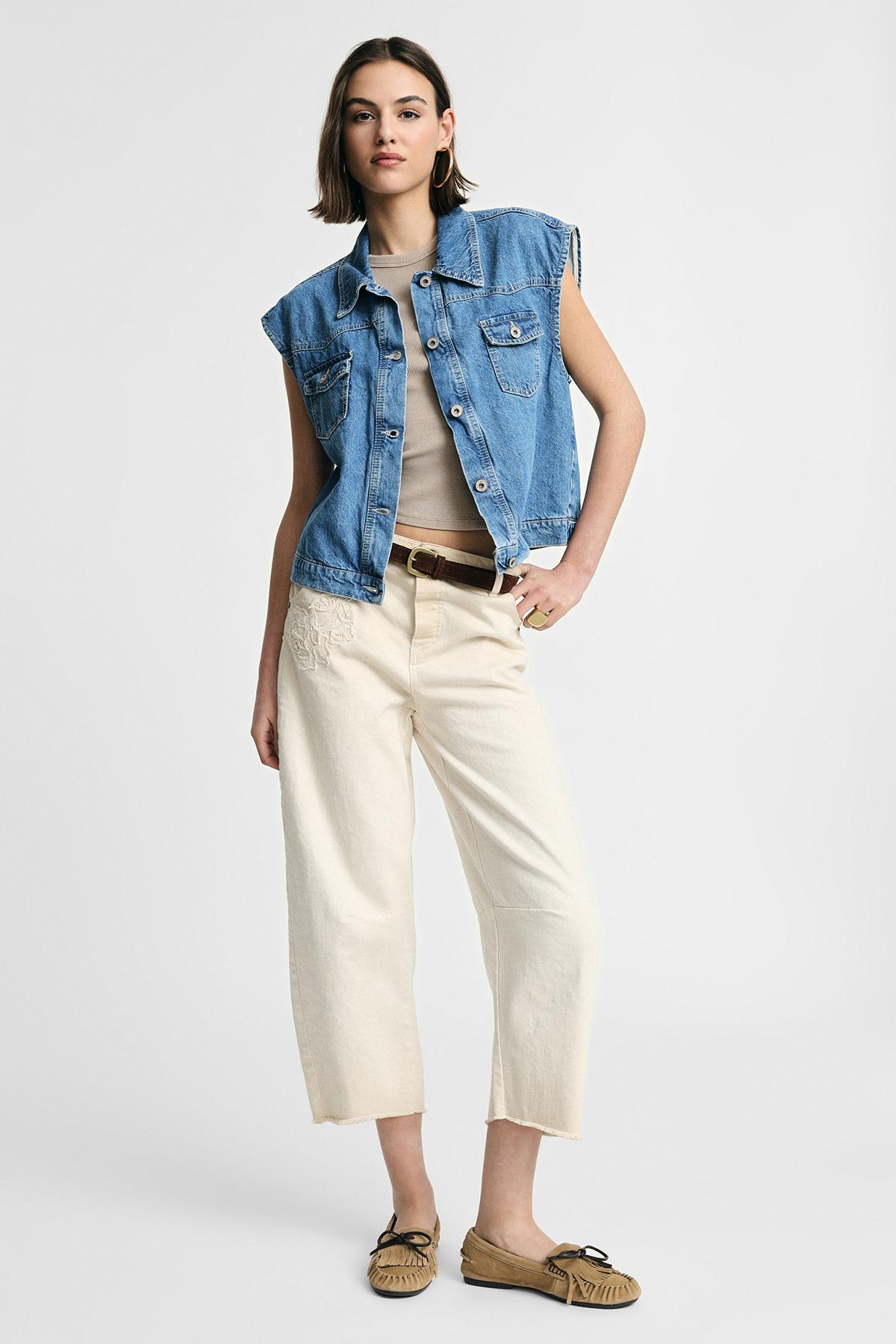 Woman wearing sleeveless denim vest, beige cropped trousers and moccasin shoes, standing with hand on hip on plain white background