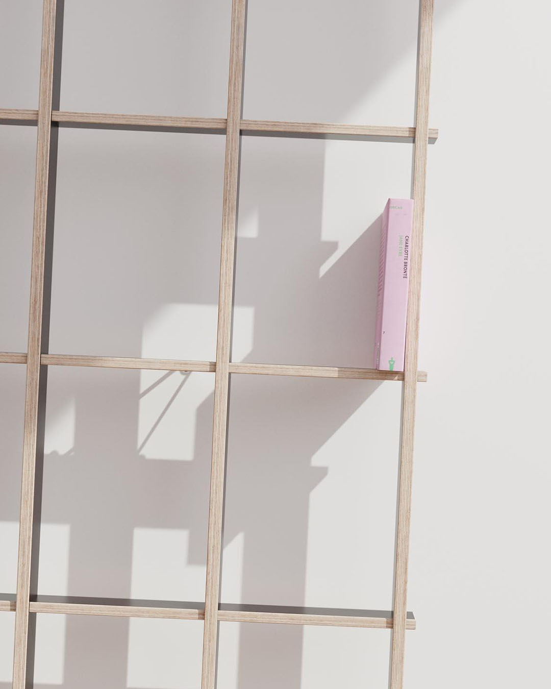 Minimalist light-wood grid shelf against a white wall casting geometric shadows, with a single vertical pink book on the right