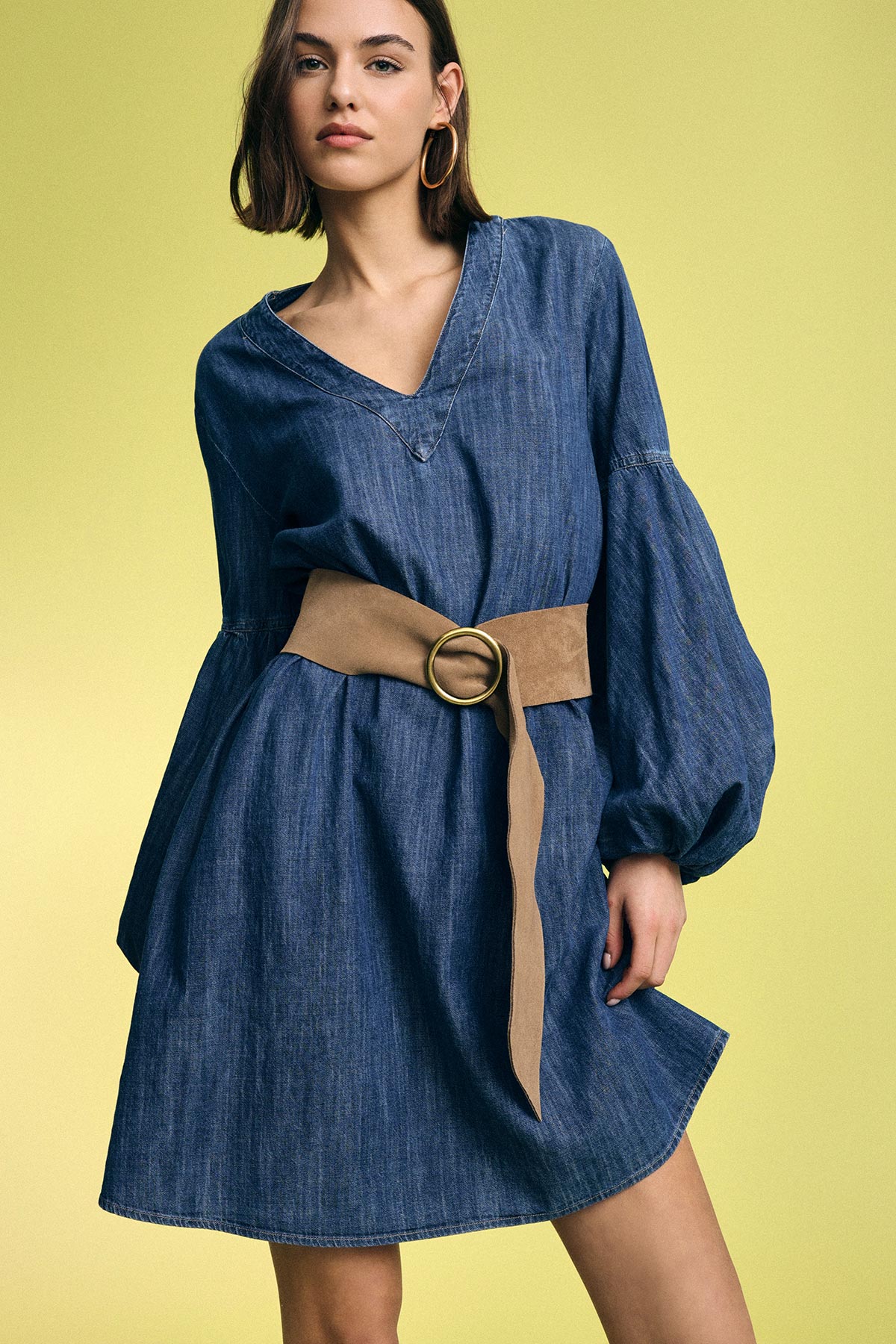 Model wearing short denim dress with voluminous sleeves and tan suede belt with round buckle, hoop earrings on pale yellow background