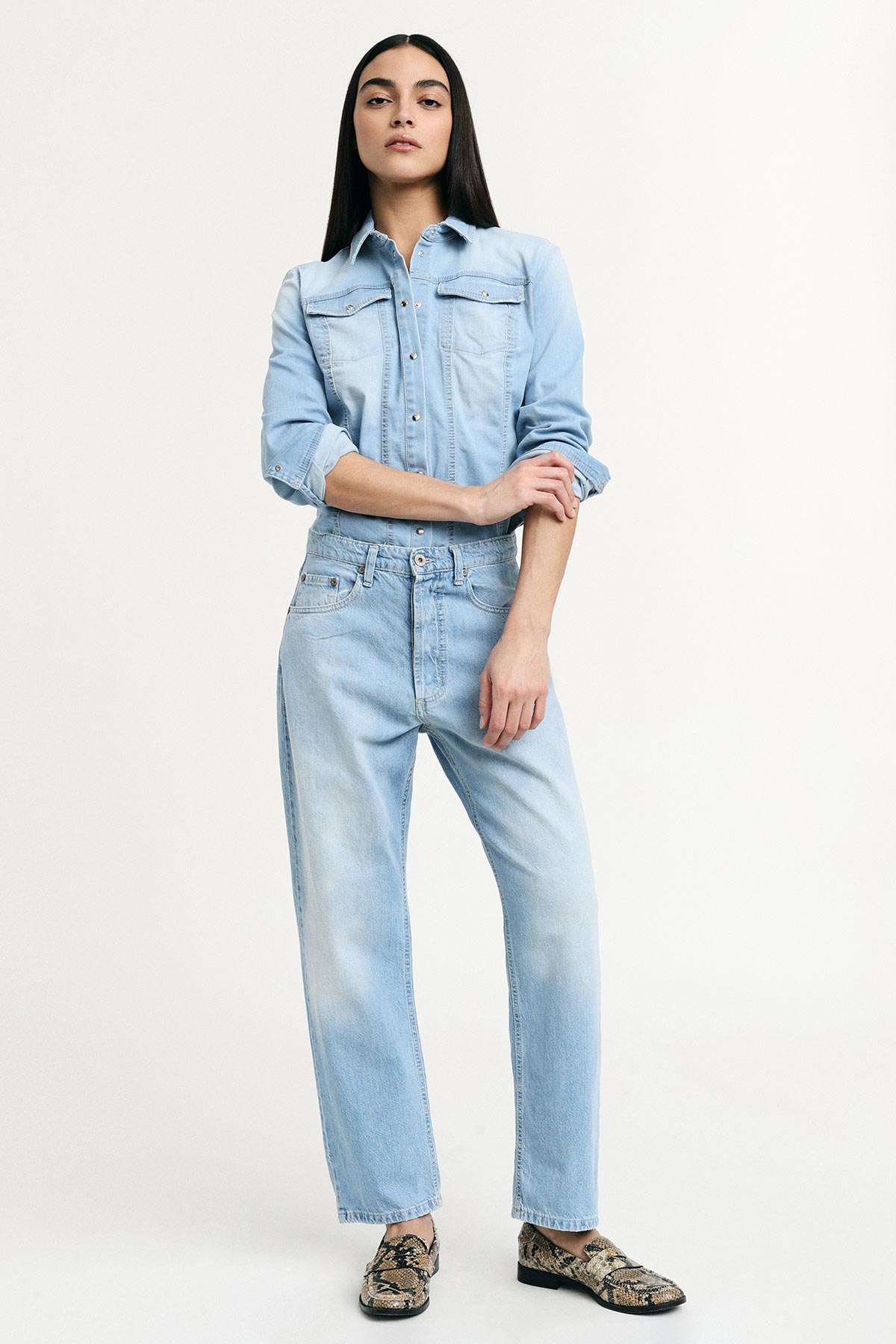 Female model wearing light denim shirt and faded high-waist jeans with snake-print loafers, standing against a white background