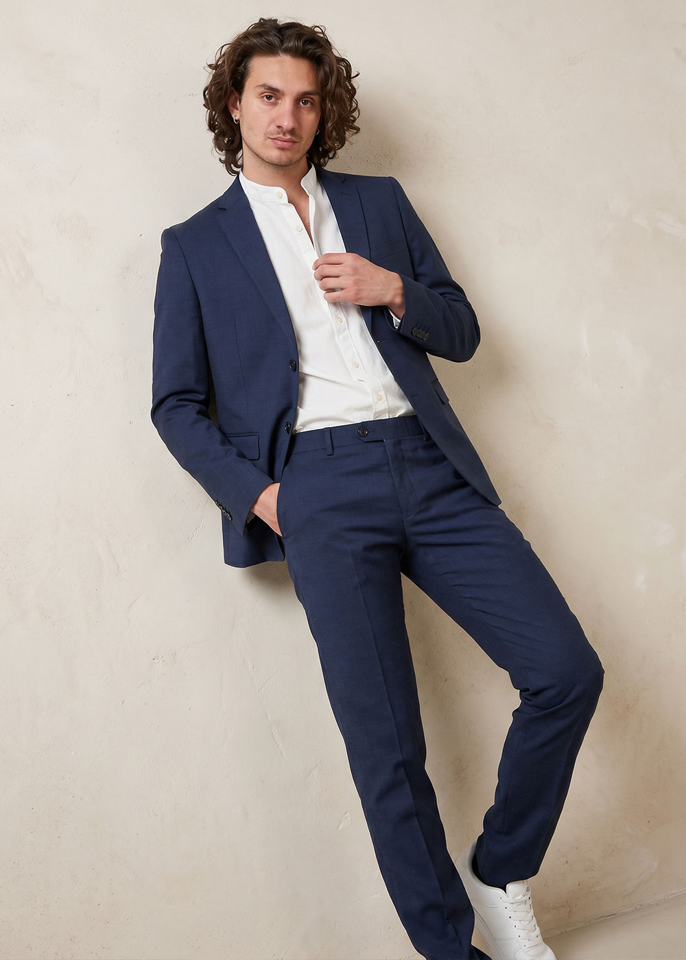 Male model wearing a navy suit, white shirt and white trainers leaning against a cream wall in a smart-casual pose