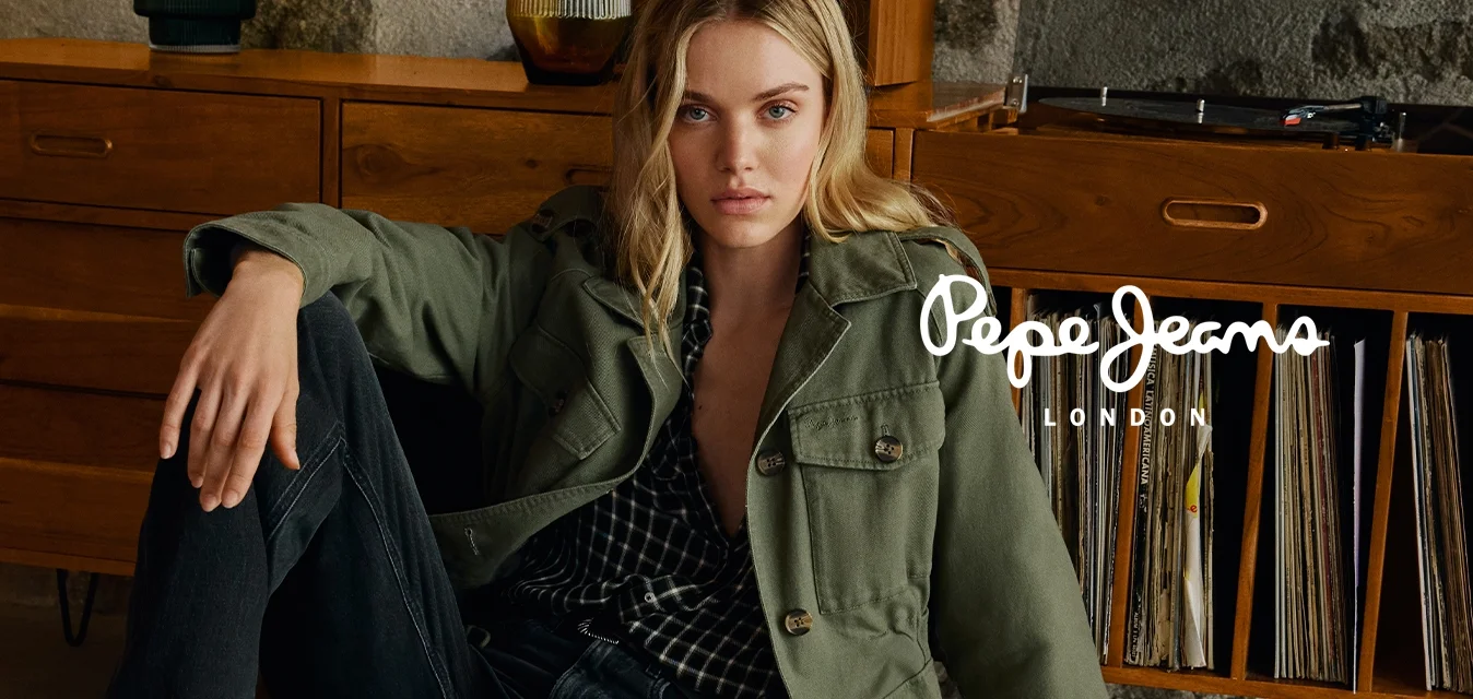 Donna bionda con giacca verde, jeans neri e stivali, seduta a terra appoggiata a un mobile in legno con dischi in vinile e vasi decorativi.