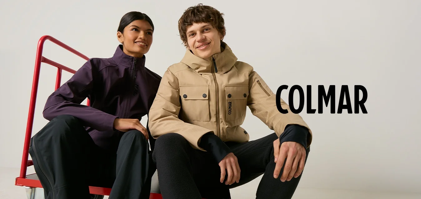 Due giovani adulti siedono su un carrello rosso, indossano giacche casual da esterno e pantaloni neri, sorridendo davanti a uno sfondo semplice.