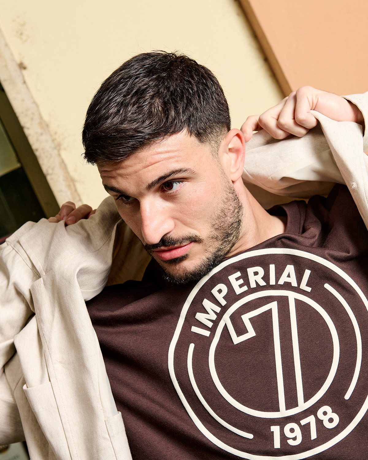 Young man with short dark hair putting on a beige jacket, wearing brown t-shirt with circular “Imperial 1978” logo