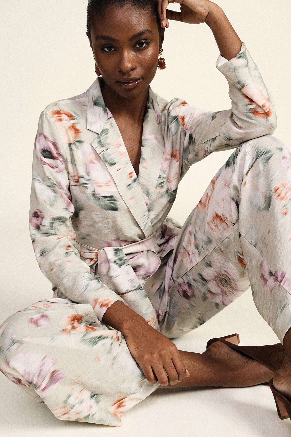 Woman wearing cream floral wrap blazer and matching trousers, seated pose highlighting statement earrings and brown heels