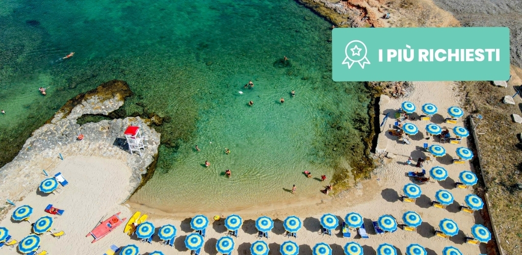 Aerial view of turquoise sea and sandy beach with rows of blue-and-white parasols, swimmers and a rocky cove