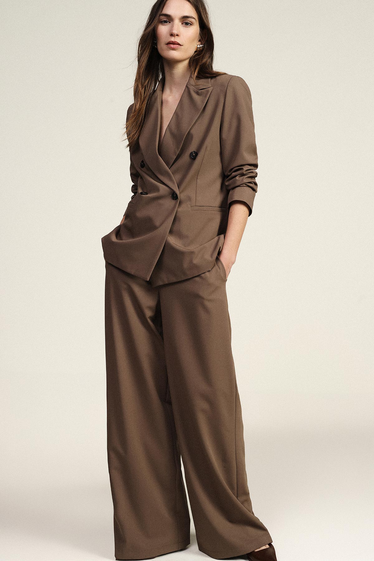 Young woman modelling a brown double-breasted blazer and wide-leg trousers, hands in pockets against a neutral studio background