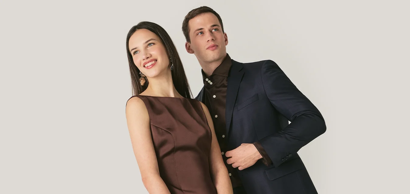 Abito di raso marrone con corpetto aderente e gonna plissettata, abbinato a un completo scuro. Sono visibili una pochette nera e un bracciale a polso metallico.