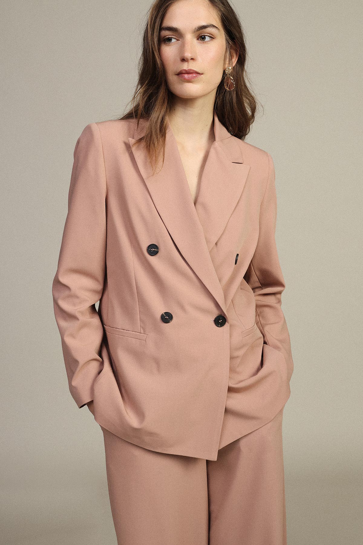 Woman wearing dusty pink double-breasted blazer and matching wide-leg trousers, hands in pockets against a neutral studio background