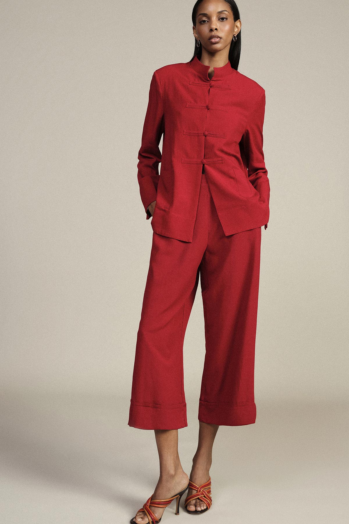 Woman in red mandarin-collar jacket and matching wide-leg cropped trousers with strappy heeled sandals on a neutral background