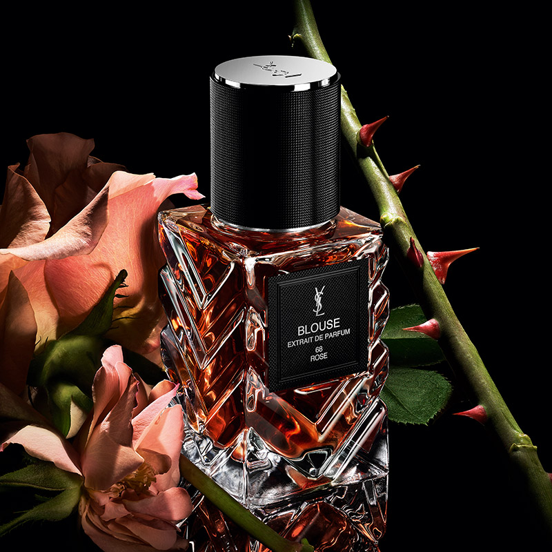 Un flacone di YSL Blouse Extrait de Parfum tra rose e uno stelo spinoso su sfondo scuro.
