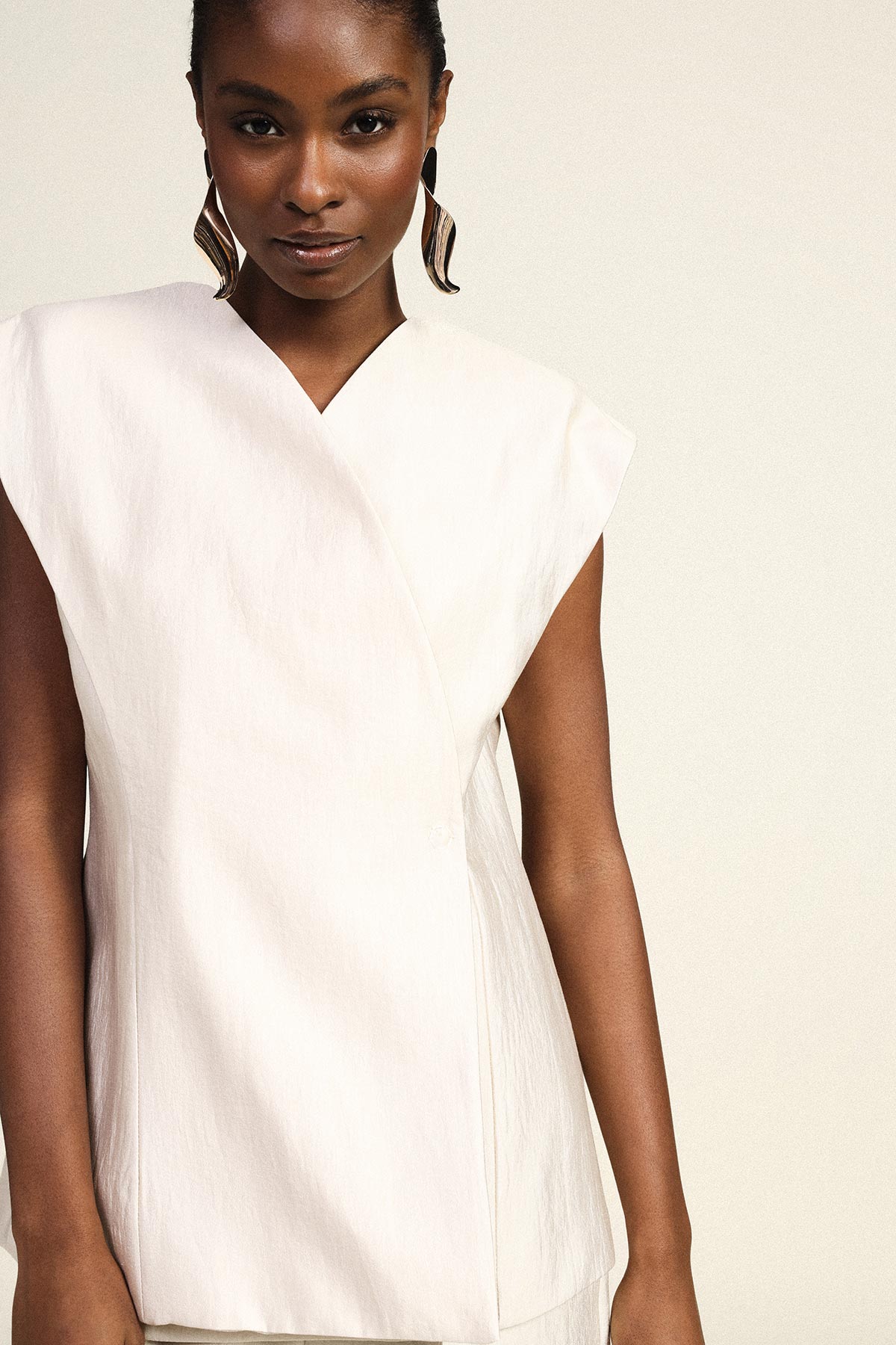 Female model wearing a sleeveless white wrap top with subtle texture and statement gold earrings, neutral background, torso view