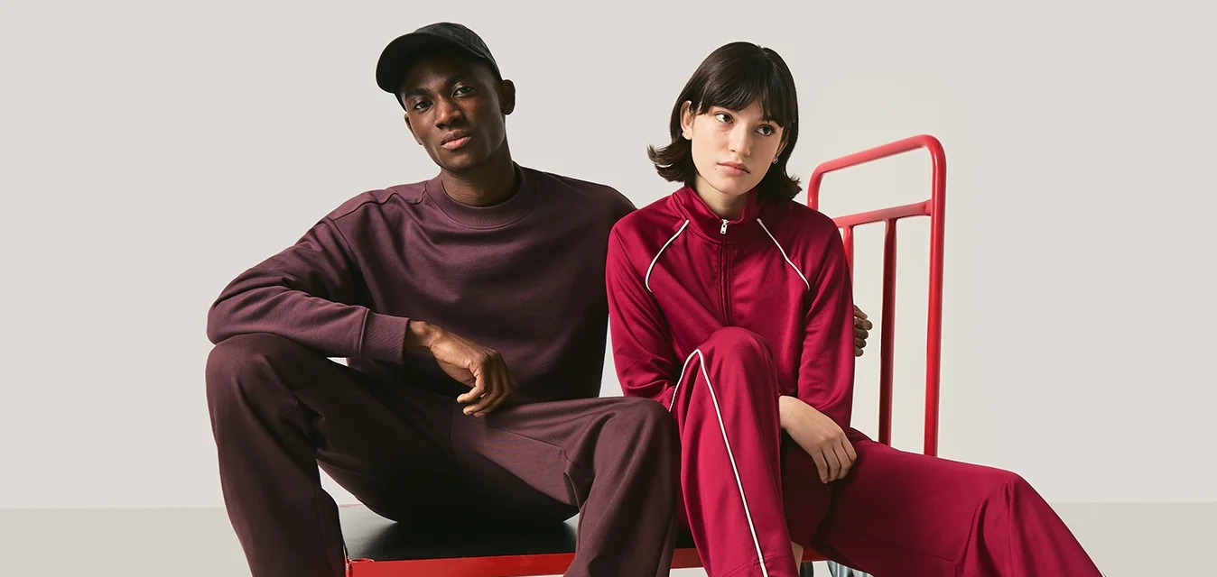 Due persone vestite in abiti casual abbinati color bordeaux sono sedute su e accanto a un carrello a piattaforma rosso, davanti a uno sfondo neutro.