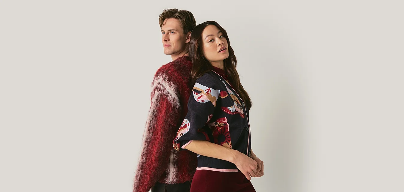 Uomo e donna in piedi schiena contro schiena davanti a uno sfondo uniforme, indossando maglioni con motivi e guardando in direzioni opposte.
