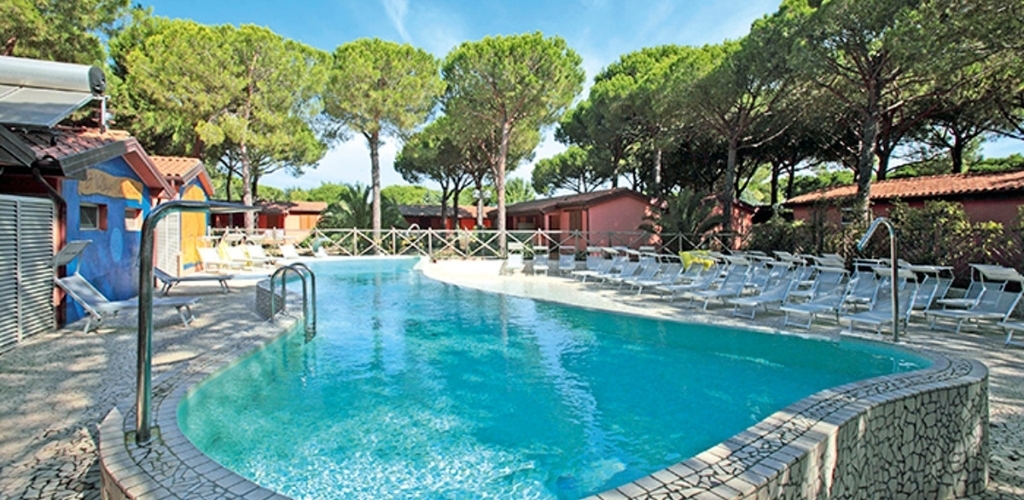 Outdoor resort swimming pool with turquoise water, rows of sun loungers, red bungalows and tall pine trees under a clear blue sky