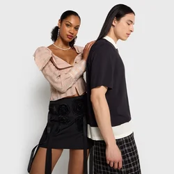 Due modelli stanno schiena contro schiena. La donna indossa un top testurizzato rosa chiaro e una gonna nera con applicazioni floreali. L'uomo indossa una camicia nera e pantaloni a quadri.