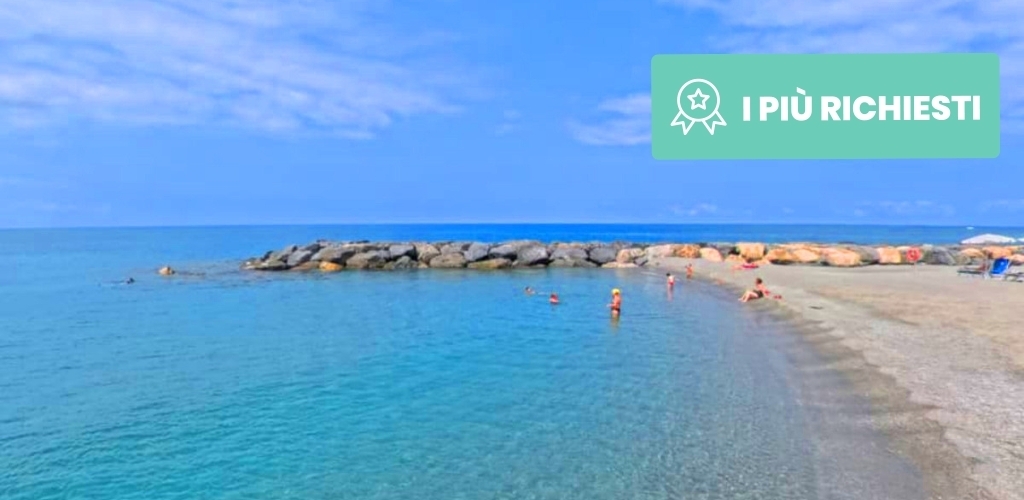 Sunny Mediterranean beach with clear turquoise water, swimmers by a rocky breakwater and pebbled shore, banner reading 'I PIÙ RICHIESTI'