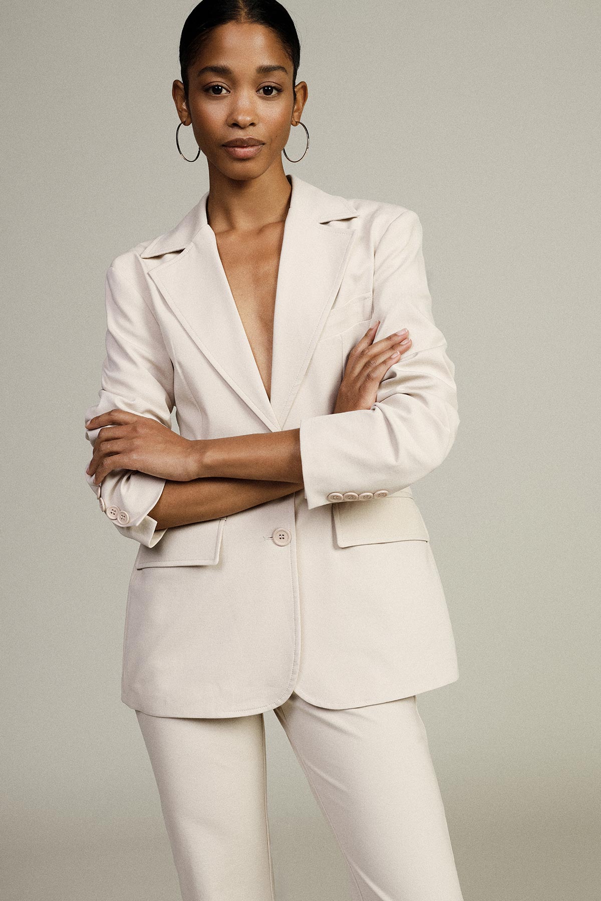 Woman in a cream tailored blazer and matching trousers with hoop earrings, arms crossed against a neutral background
