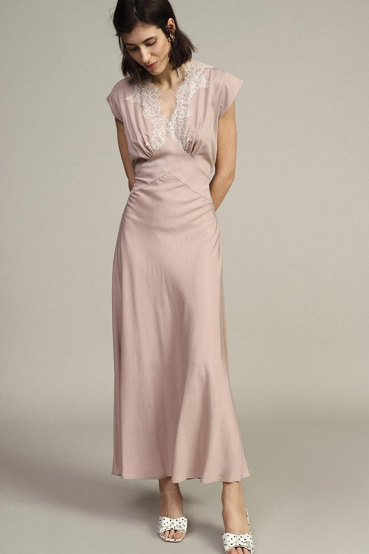 Woman in a blush pink lace-trimmed maxi dress and white heeled sandals, looking down in studio