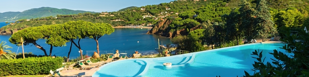 Infinity pool with sun loungers overlooking a Mediterranean bay framed by umbrella pines and coastal hills