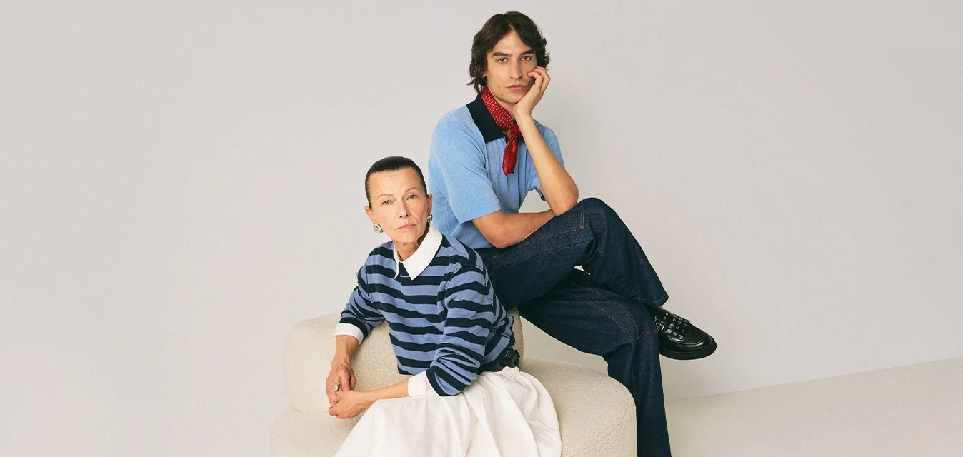 Due persone sedute su una sedia rotonda beige contro uno sfondo semplice, indossando abbigliamento casual tra cui stivali e maglioni.