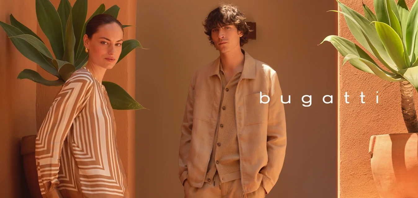 Due persone con abiti in tonalità neutre sono in piedi su scalini in terracotta vicino a piante in vaso, con colline rocciose e cielo nuvoloso sullo sfondo.