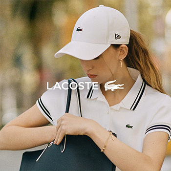 Woman wearing white Lacoste polo shirt and cap adjusting a navy tote bag in a blurred outdoor setting with visible Lacoste logo