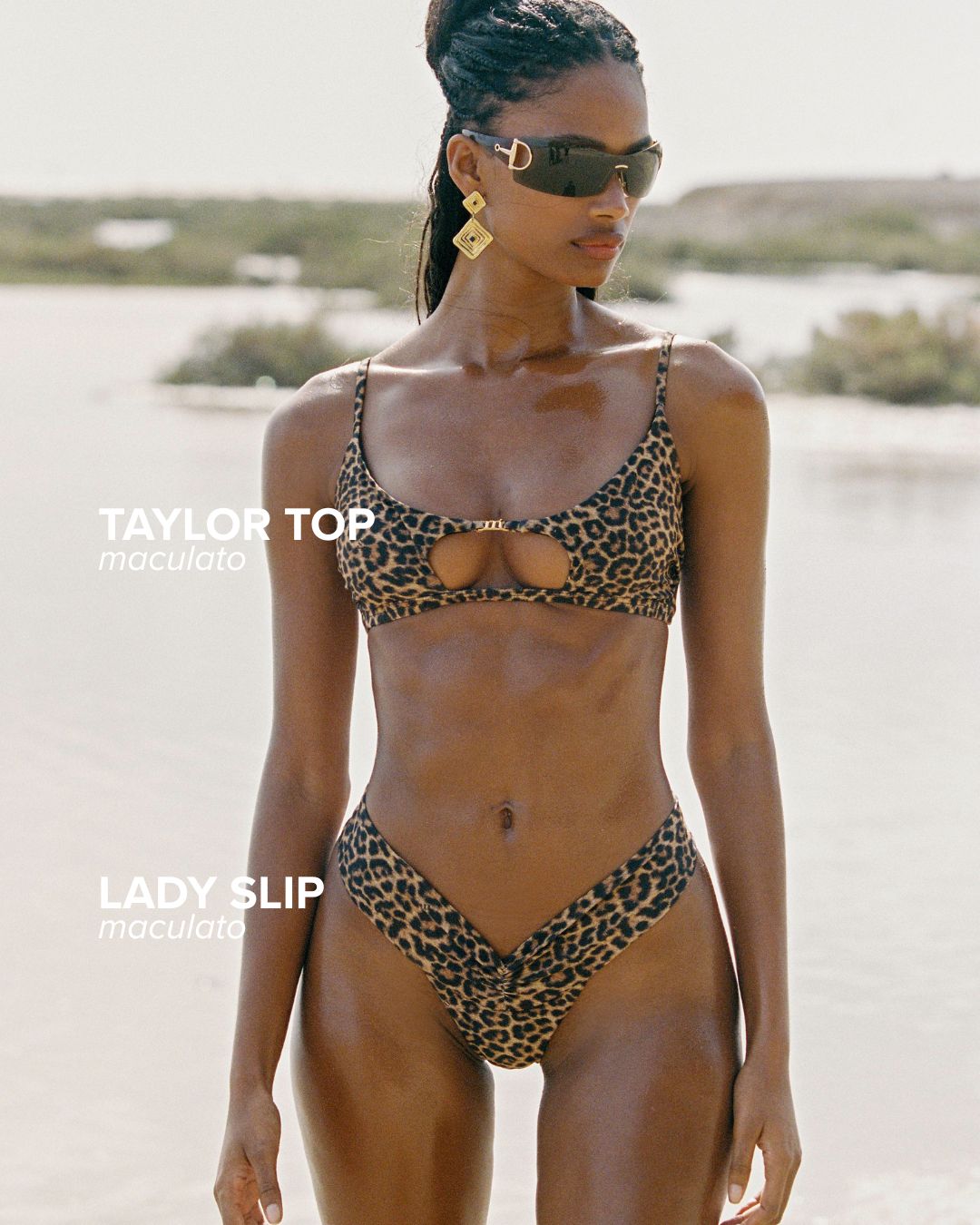 Woman in leopard-print bikini and sunglasses with gold geometric earrings standing on a sunlit beach near shallow water